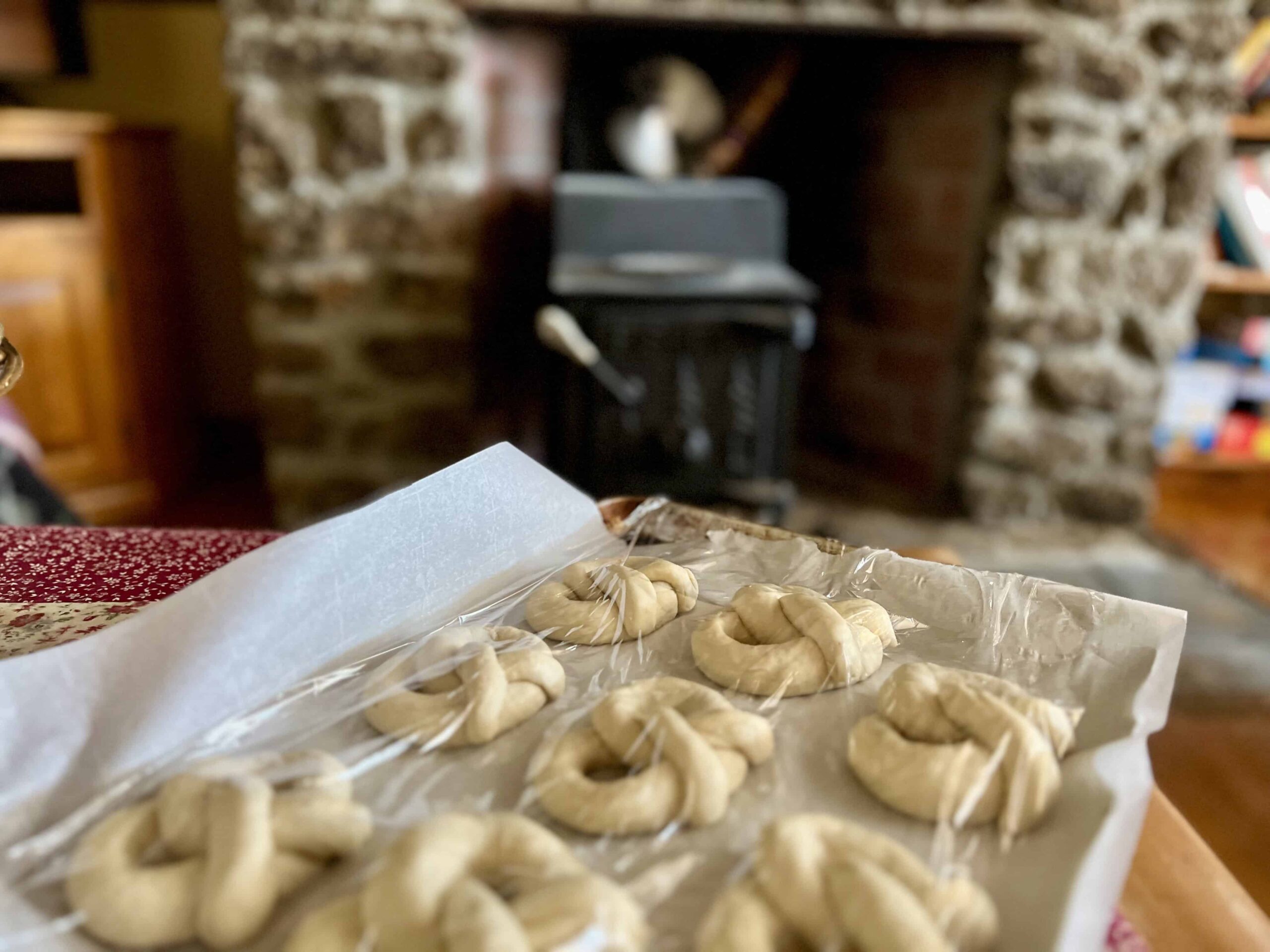 Shaped sourdough soft pretzels covered with plastic wrap and rising on a tray near a warm stone fireplace in a cozy kitchen setting.