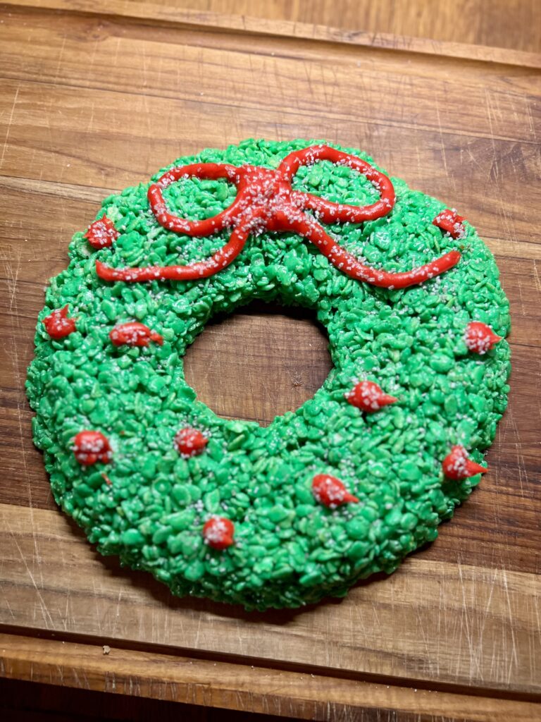 Large Christmas Rice Krispie wreath decorated with a red bow and red berries, shaped into a festive holiday wreath on a wooden cutting board.
