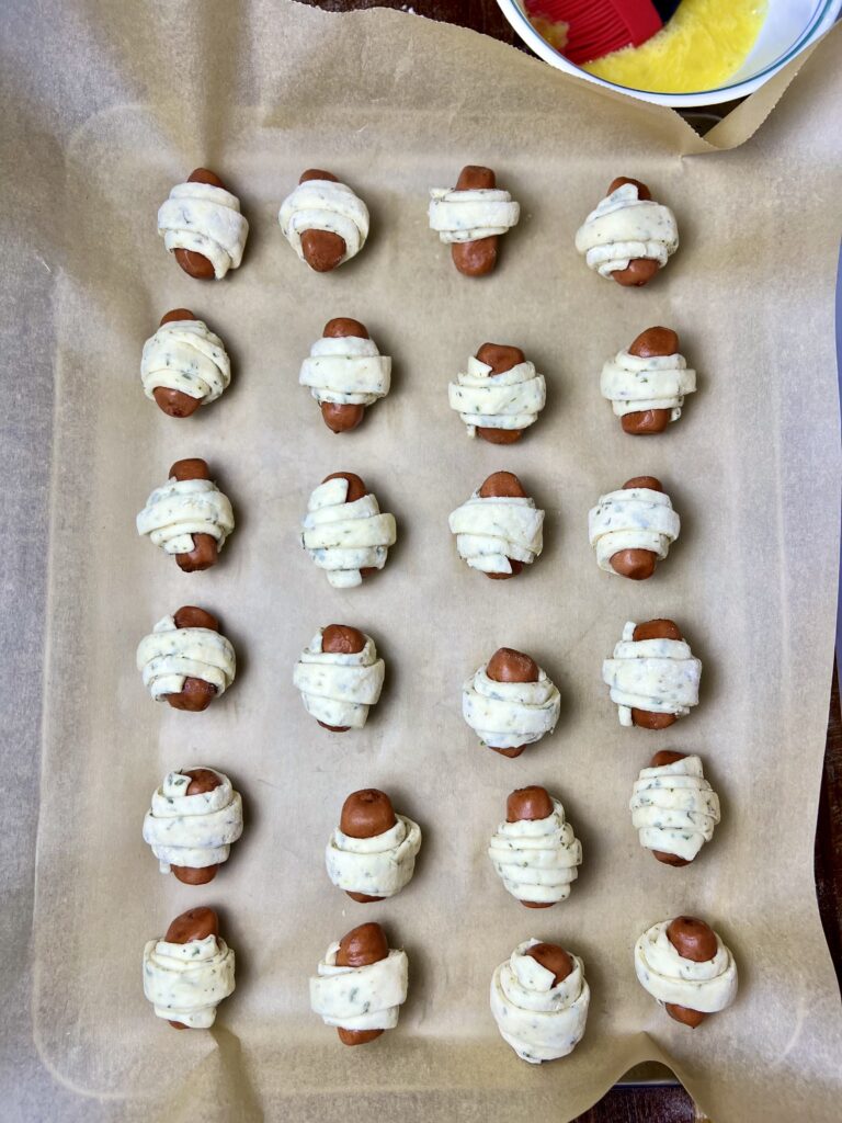 Unbaked pigs in a blanket arranged on a parchment-lined baking sheet, showing mini sausages wrapped in herbed homemade dough before baking.