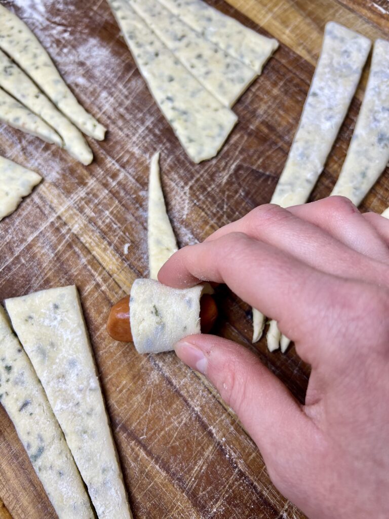 Hand rolling herbed dough around a mini sausage on a floured wooden cutting board while assembling homemade pigs in a blanket.