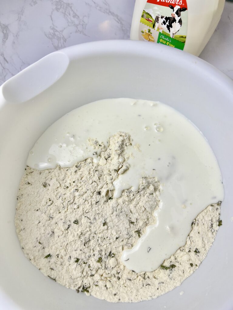 Buttermilk being poured into a bowl of seasoned flour mixture to make homemade pigs in a blanket dough, showing the step of combining wet and dry ingredients.