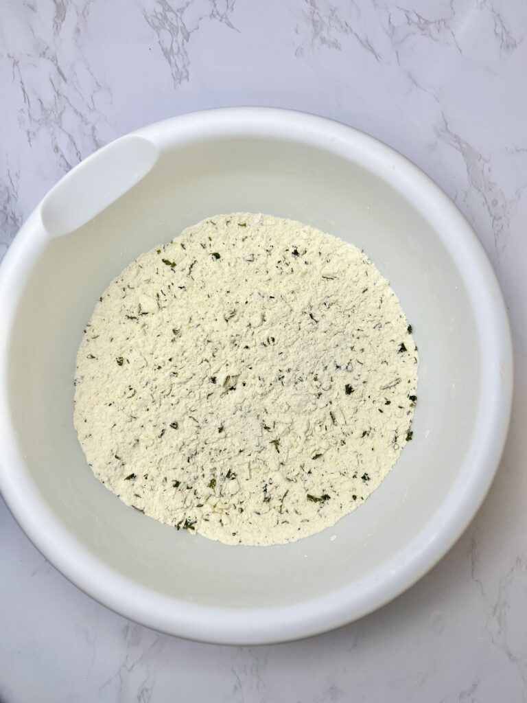 Dry ingredients for homemade pigs in a blanket dough mixed in a white bowl, showing flour blended with herbs and seasonings on a marble countertop.