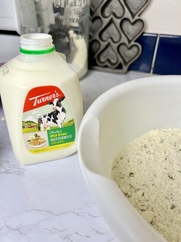 Buttermilk next to a bowl of seasoned flour mixture, showing ingredients being prepared for homemade pigs in a blanket dough on a kitchen counter.