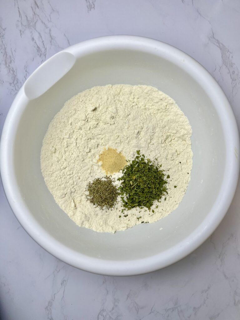 Dry ingredients and seasonings for pigs in a blanket dough in a white mixing bowl, showing flour with herbs and spices added before mixing.