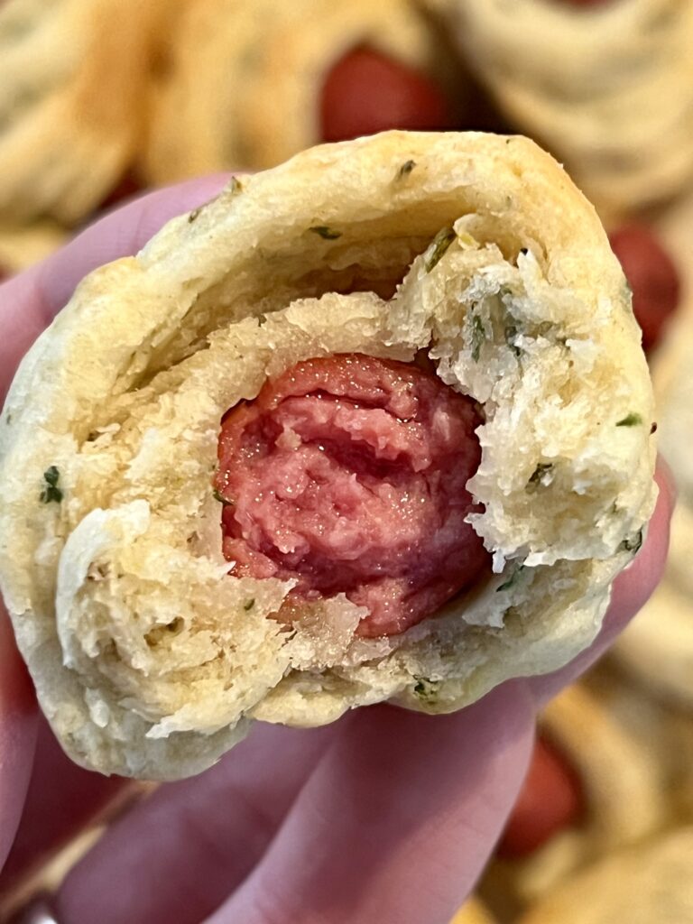 Close-up of a pig in a blanket bitten open, showing a juicy mini smoked sausage wrapped in soft, fluffy homemade dough with visible herb flecks.