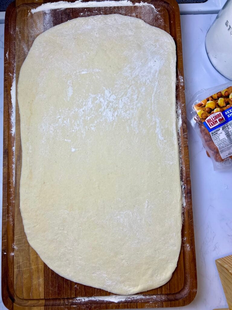 the dough rolled out on a wooden cutting board, ready to be cut into the shapes for making the pigs in a blanket. 