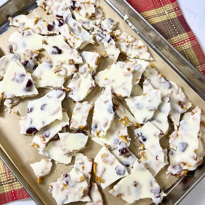 White chocolate bark setting on a baking sheet with pretzels, dried cranberries, and chopped nuts evenly distributed