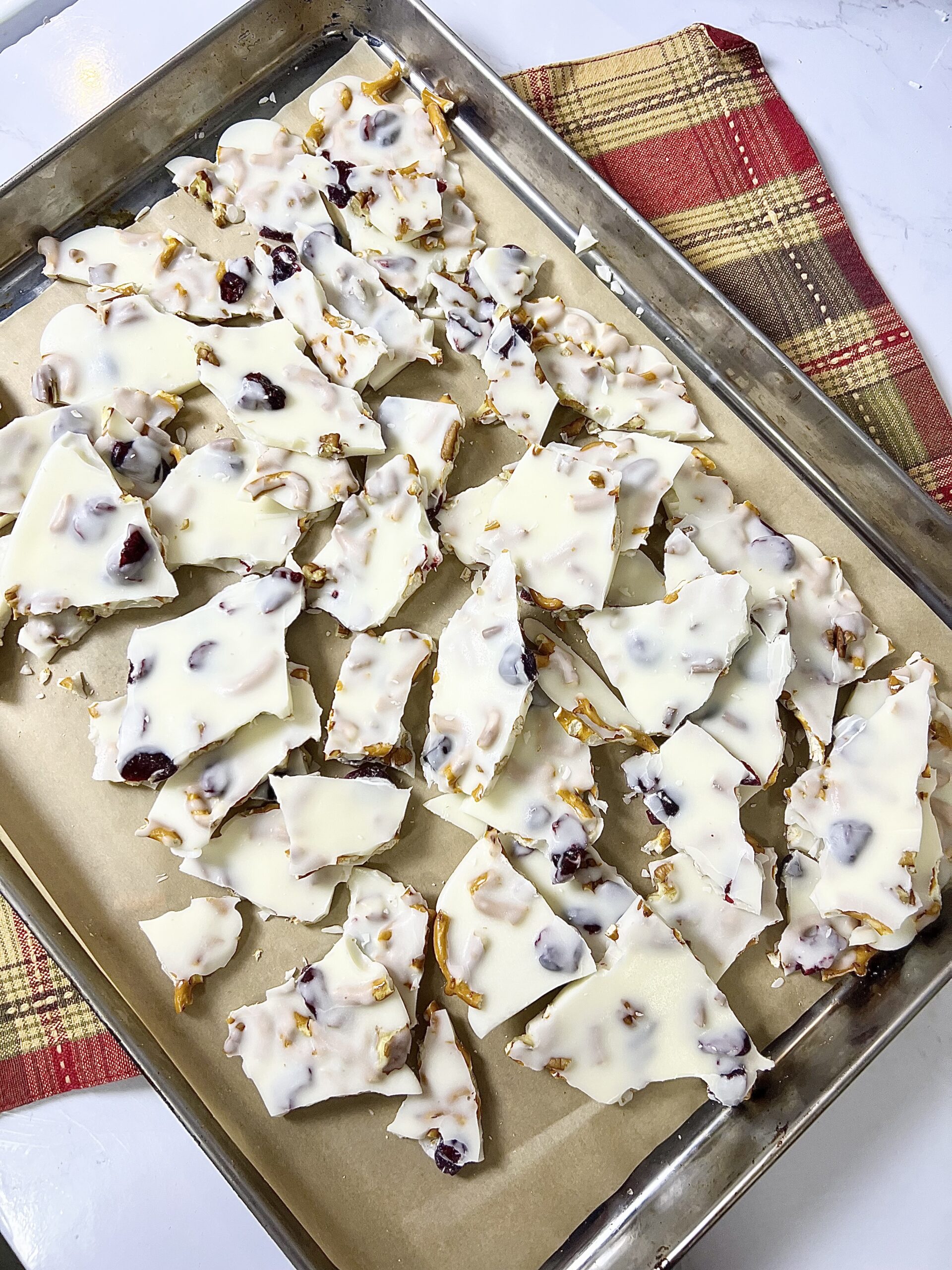 White chocolate bark setting on a baking sheet with pretzels, dried cranberries, and chopped nuts evenly distributed