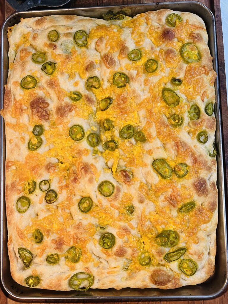 Golden baked jalapeño cheddar sourdough focaccia in a sheet pan with melted cheese, blistered crust, and sliced jalapeños.