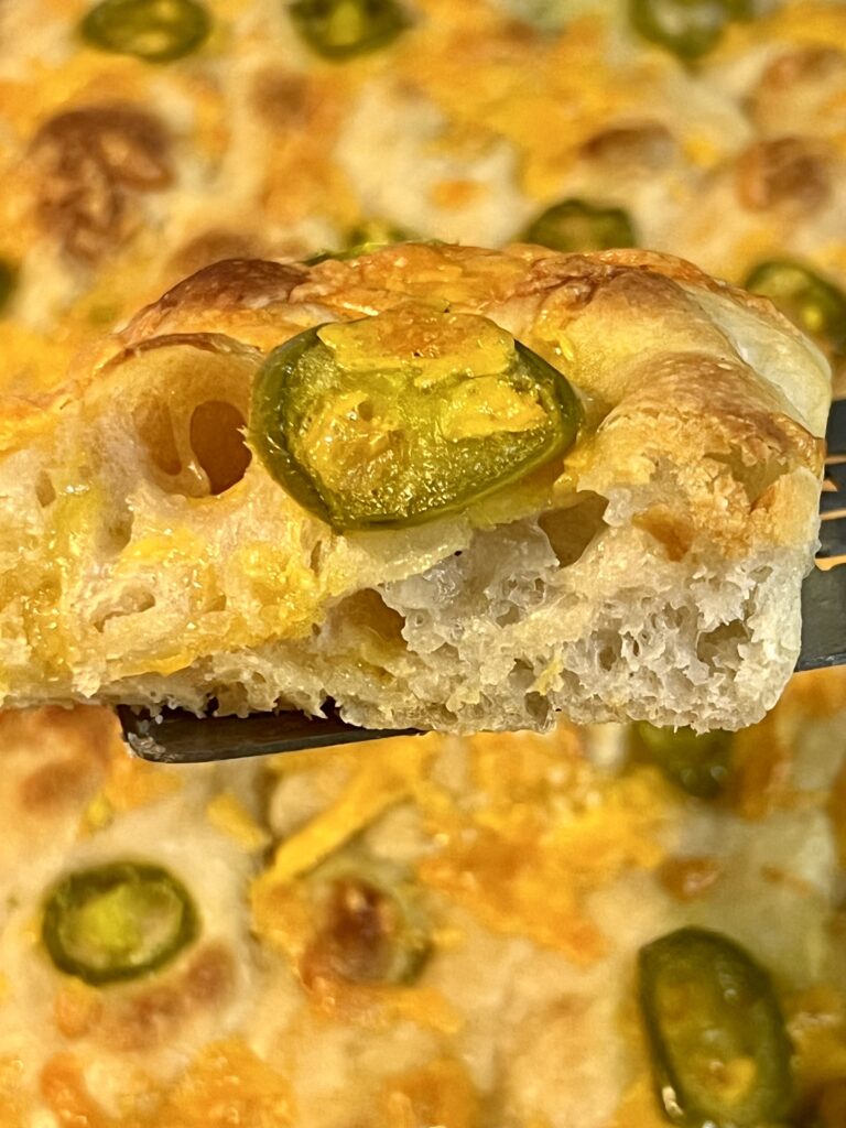 Close-up of a slice of jalapeño cheddar sourdough focaccia lifted from the pan, highlighting soft interior crumb and melted cheddar.