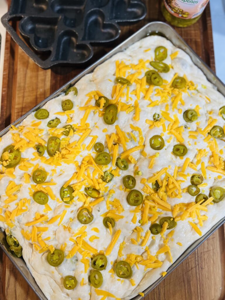 Jalapeño cheddar sourdough focaccia dough in an oiled baking pan topped with sliced jalapeños and shredded sharp cheddar before baking.