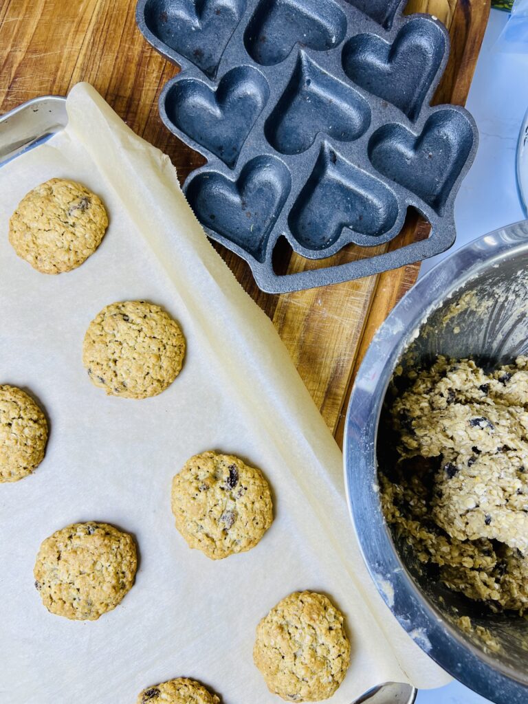 Baked oatmeal raisin cookies cooling on parchment paper next to mixing bowl and heart shaped baking pan