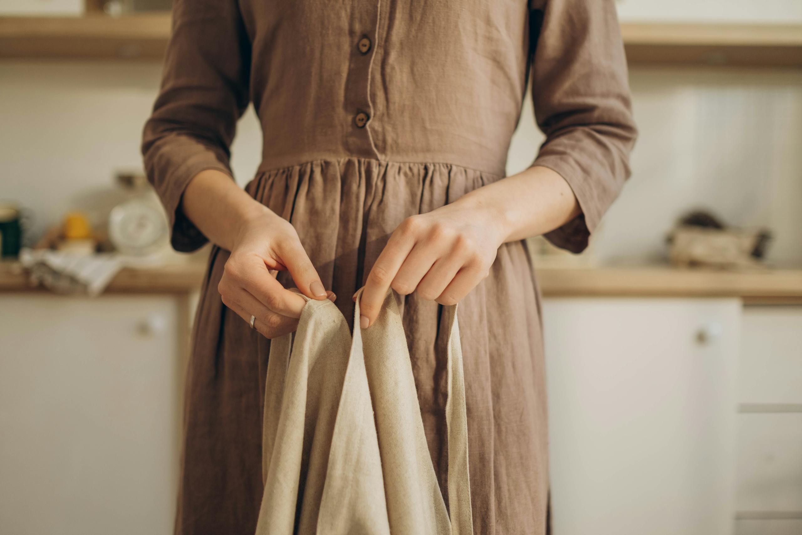 Close-up of a woman adjusting her apron in a rustic kitchen setting, wearing a brown dress.