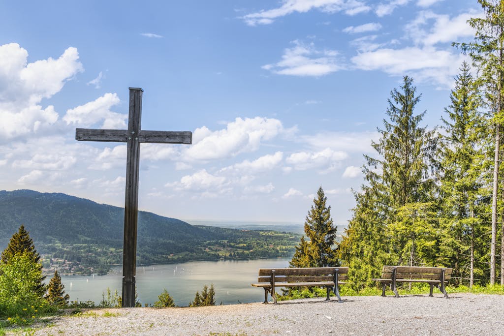 Serene landscape of Tegernsee in Germany featuring wooden cross, benches, and lush trees under a blue sky.