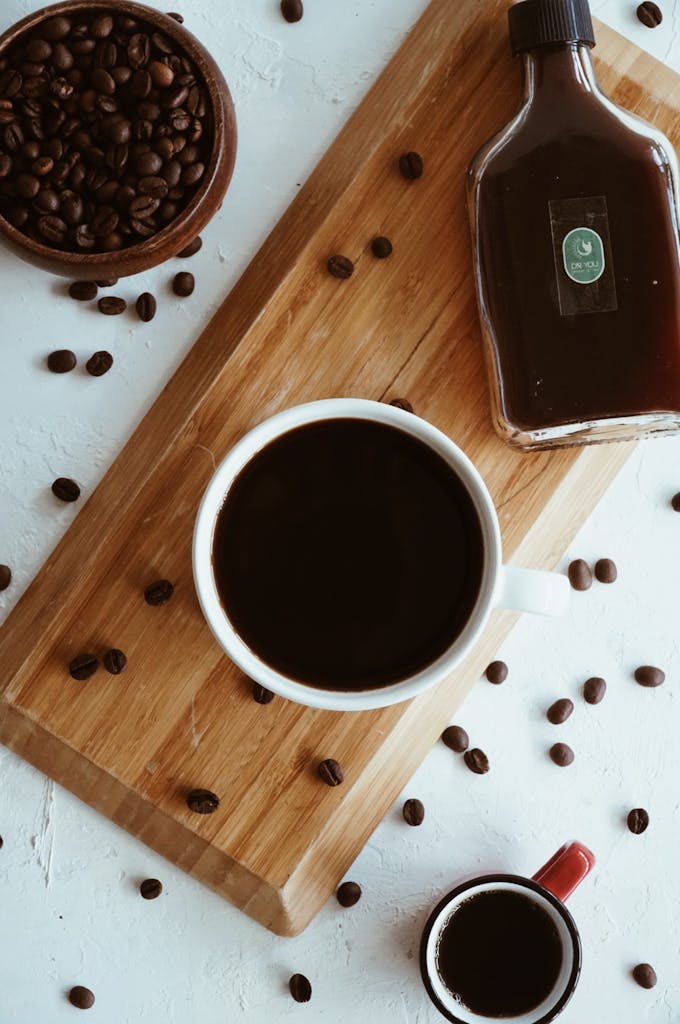 Coffee cups and a syrup bottle on a wooden board with scattered beans. Perfect for food photography.