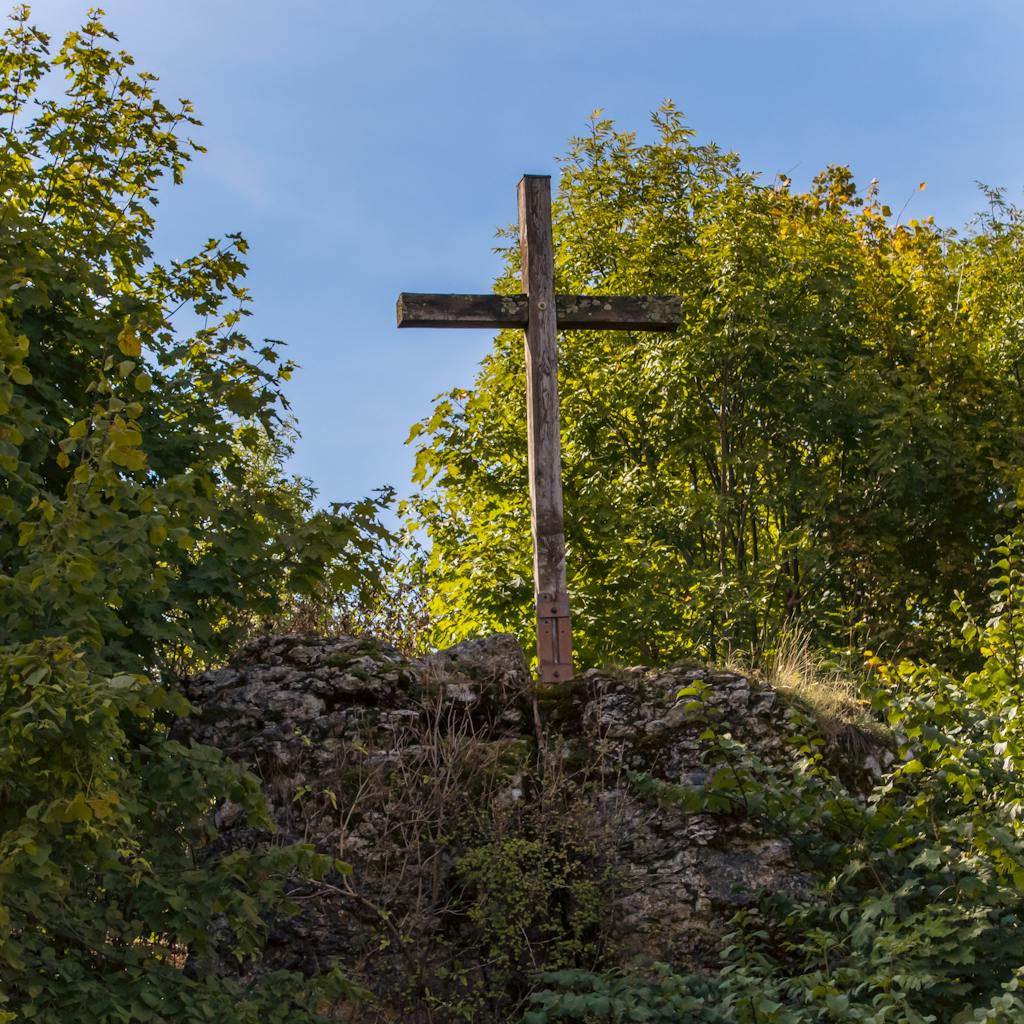 Rustic wooden cross on a rocky cliff surrounded by trees under a bright blue sky.