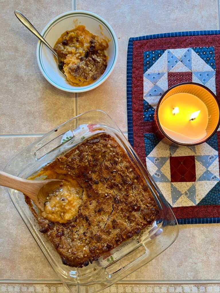 Homemade sweet potato casserole with canned yams baked in a glass dish with a buttery brown sugar pecan topping, served with a spoonful in a bowl and a lit candle beside it.