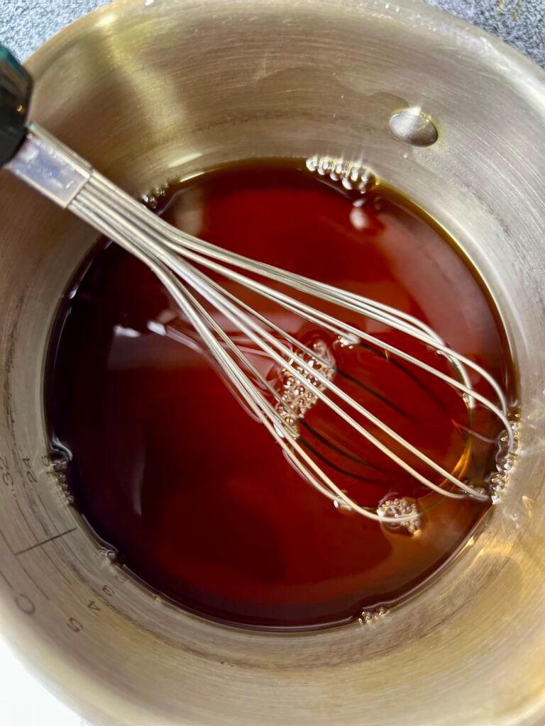 Homemade coffee syrup simmering in a saucepan with whisk, showing rich dark brown syrup.