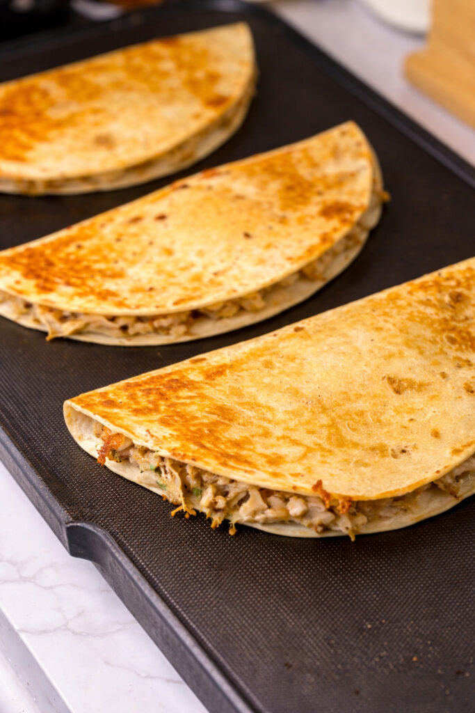Authentic Mexican chicken quesadilla recipe cooking on a large skillet with flour tortillas turning golden brown and filled with seasoned chicken and melted cheese