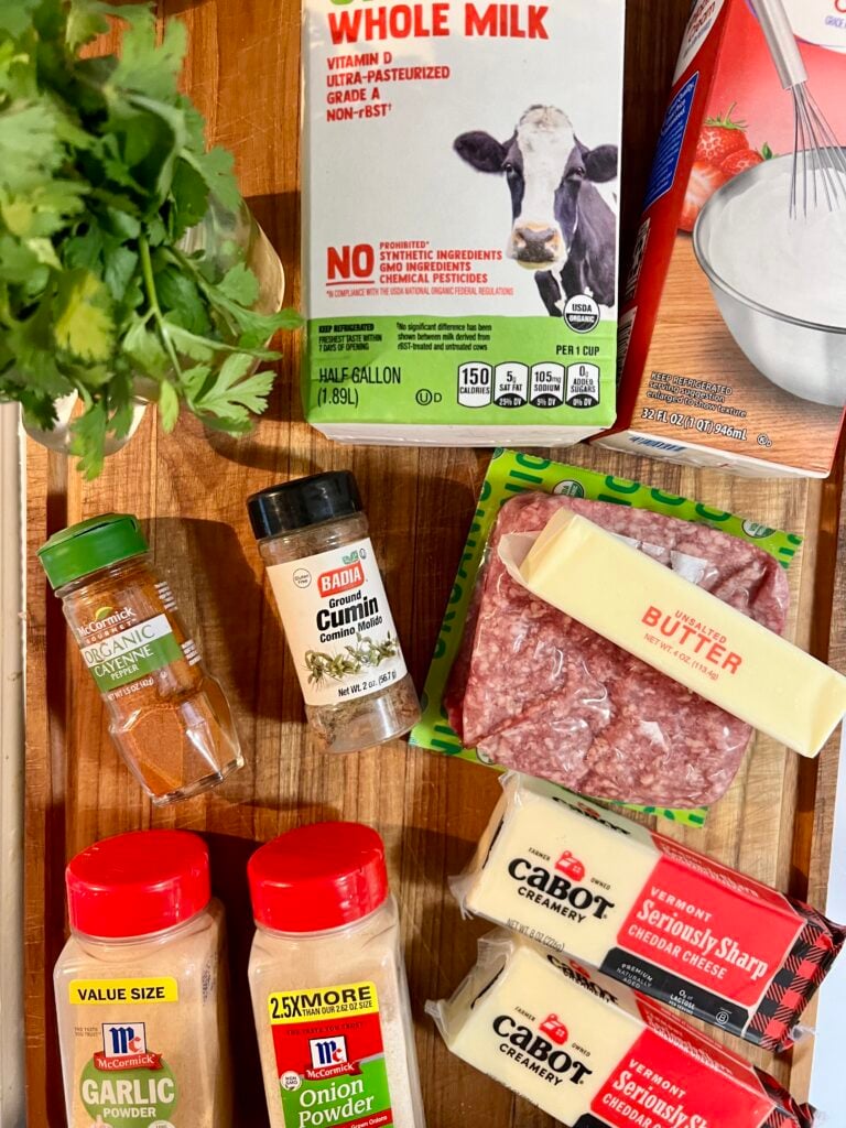 Ingredients for homemade queso dip with ground beef including cheddar cheese, milk, ground beef, butter, cilantro, and spices on a cutting board.