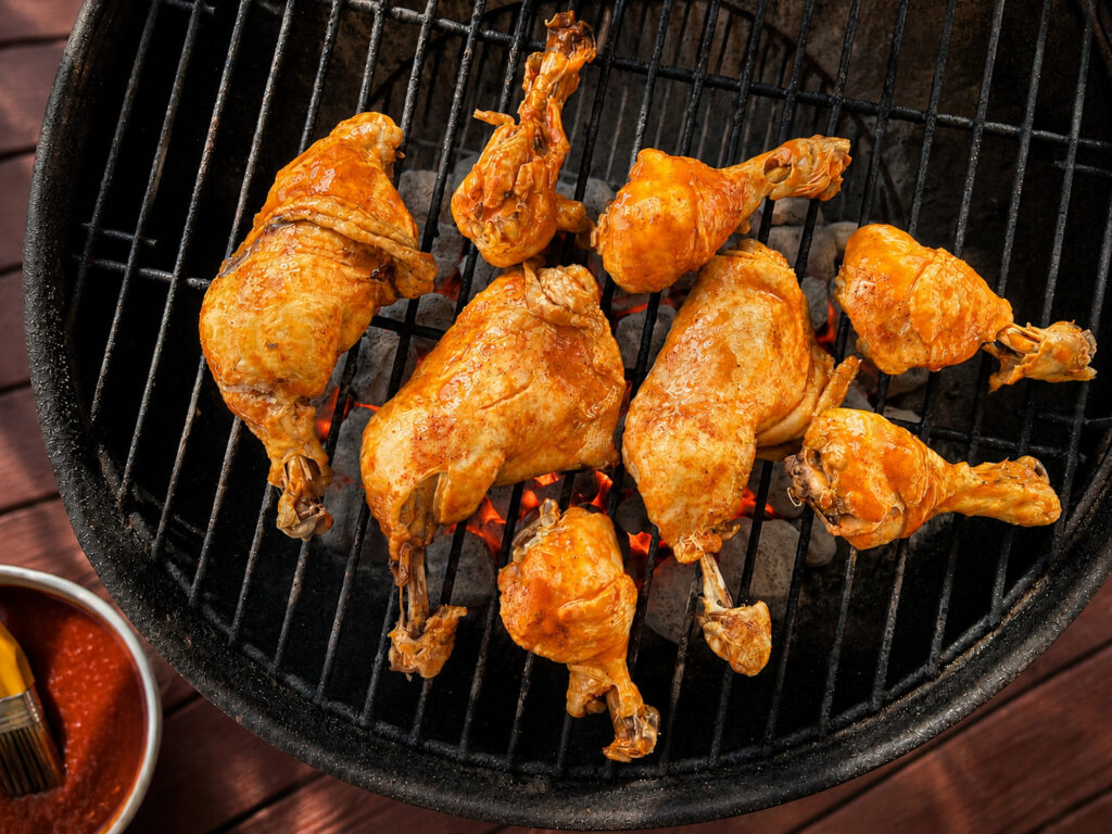 Grilled chicken drumsticks and thighs cooking over charcoal on a backyard grill, coated in a bright orange marinade or sauce, summer barbecue outdoor cooking close-up.
