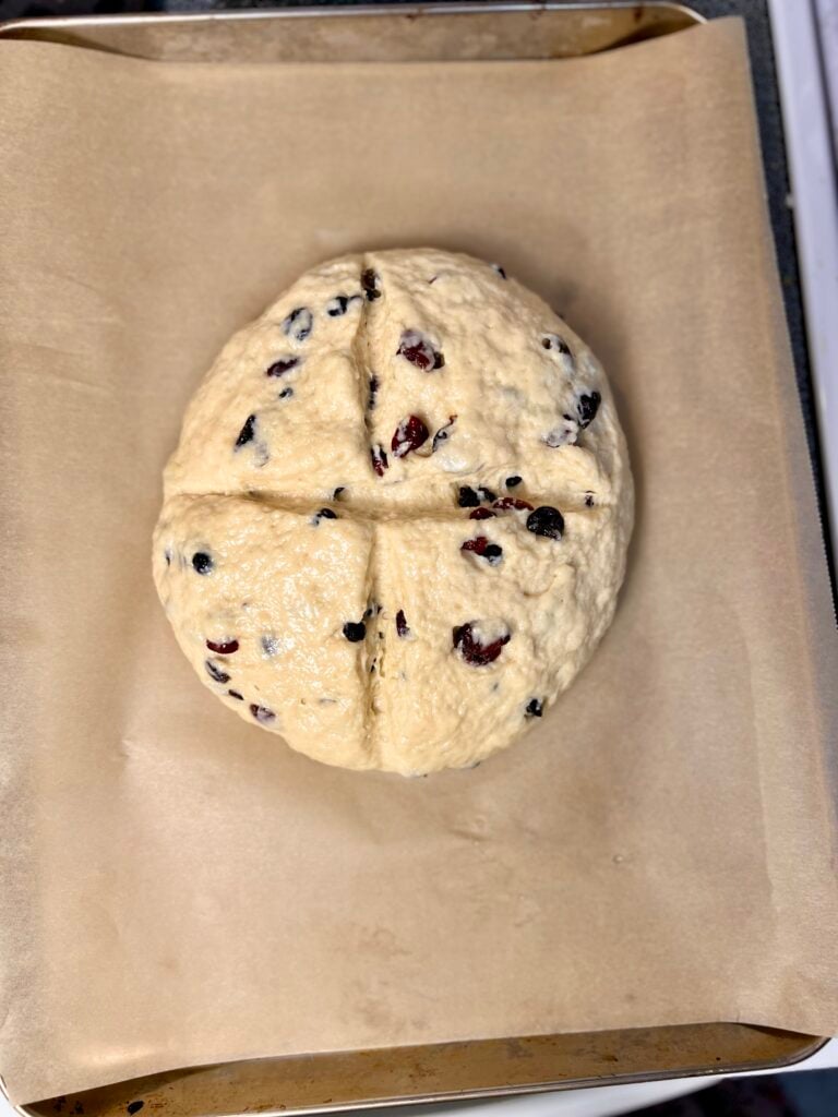 Unbaked Irish soda bread dough shaped into a round loaf with a traditional cross cut on top, studded with raisins and dried fruit on parchment-lined baking sheet.