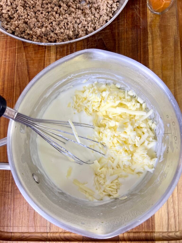 Shredded white cheese being added to a creamy queso sauce base for homemade queso dip with ground beef.