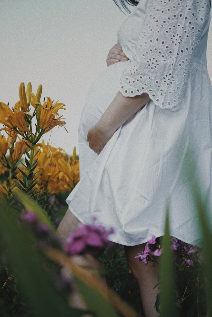 Side view of a pregnant woman in a white dress walking among vibrant flowers outdoors.