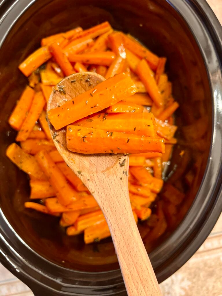 Slow cooker glazed carrots coated in buttery brown sugar glaze, tender carrot sticks lifted with wooden spoon inside crock pot.