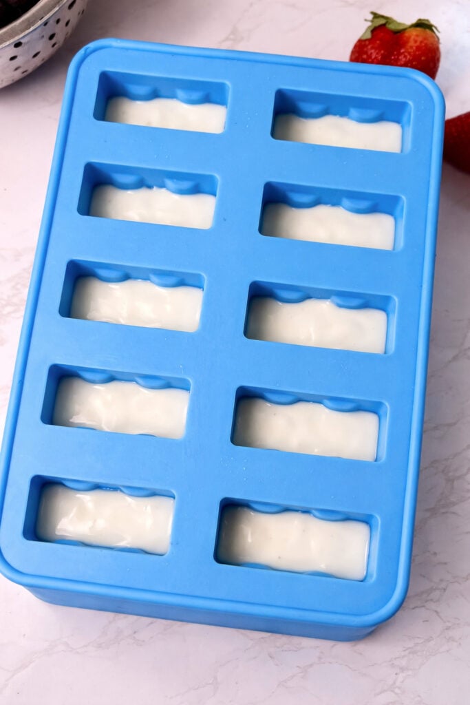 Creamy white yogurt gently fills the popsicle molds, creating a beautiful contrast against the vibrant red layer below and setting the stage for the final touch of blue.