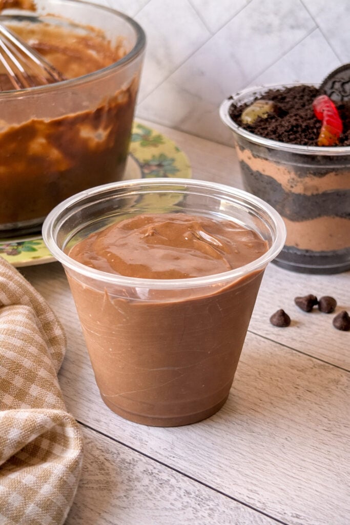 Close-up of homemade chocolate pudding in a plastic container, highlighting the thick, velvety texture with chocolate chips and layered dessert in the background
