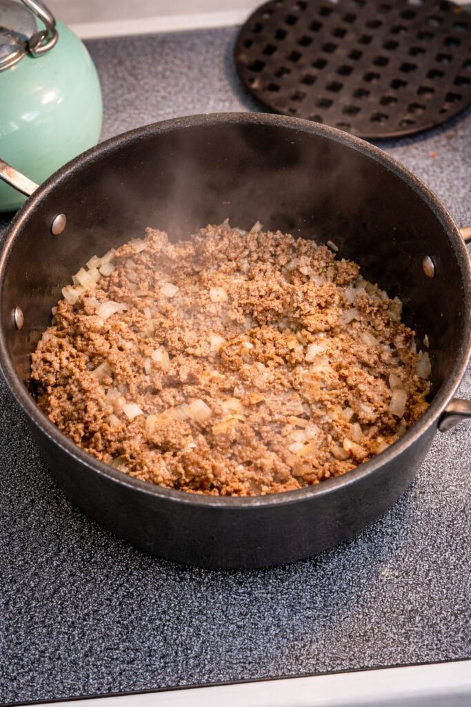 American chop suey recipe beef showing ground beef and diced onions cooking in a large pot over medium-high heat before adding sauce
