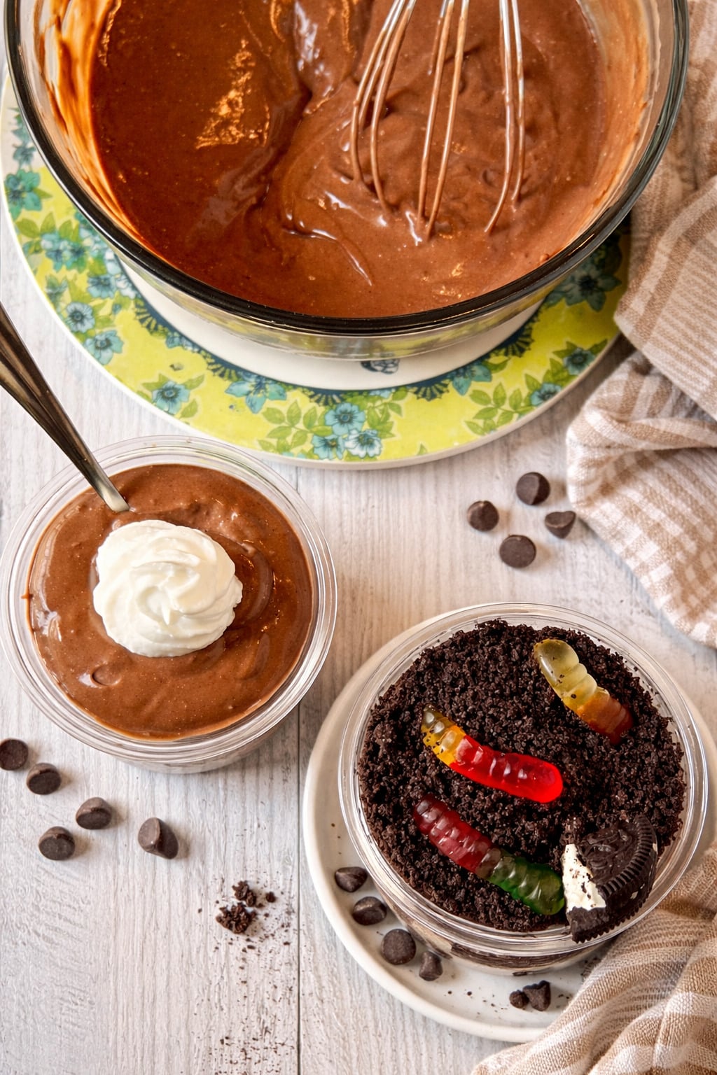 Homemade chocolate pudding being mixed in a bowl with a whisk, served with whipped cream and alongside a worms in dirt dessert with crushed cookies and gummy worms