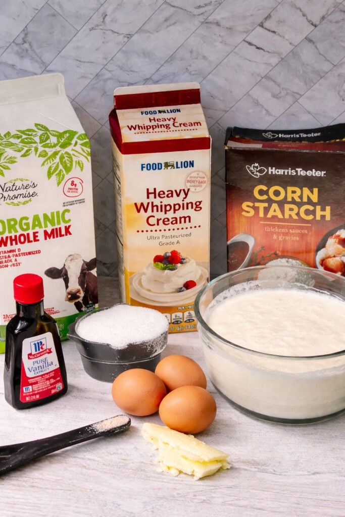 Ingredients for homemade vanilla pudding including milk, heavy cream, eggs, sugar, cornstarch, vanilla extract, and butter on kitchen counter