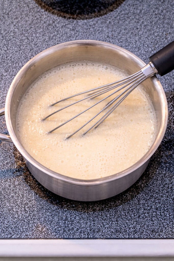 Homemade vanilla pudding cooking in a saucepan on the stovetop with a whisk, creamy mixture thickening for an easy from-scratch vanilla pudding recipe