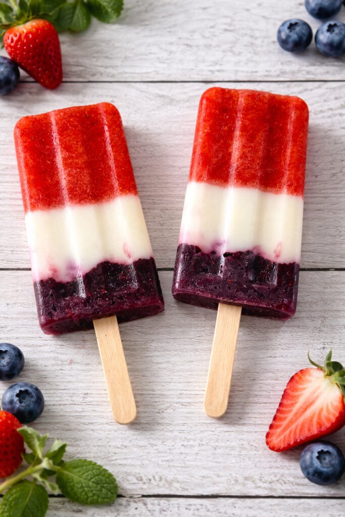 Two finished red white and blue popsicles with strawberry top, yogurt middle, and blueberry bottom on a wooden surface with fresh berries