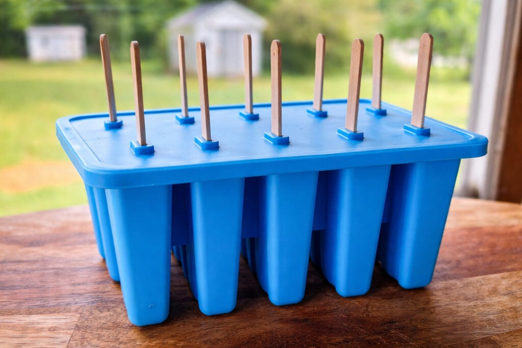Blue silicone popsicle mold with wooden sticks inserted, sitting on a wooden table outdoors with blurred backyard background