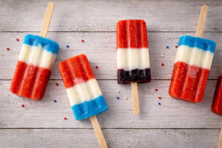 Four red white and blue popsicles laid out on a wooden table with patriotic sprinkles scattered around