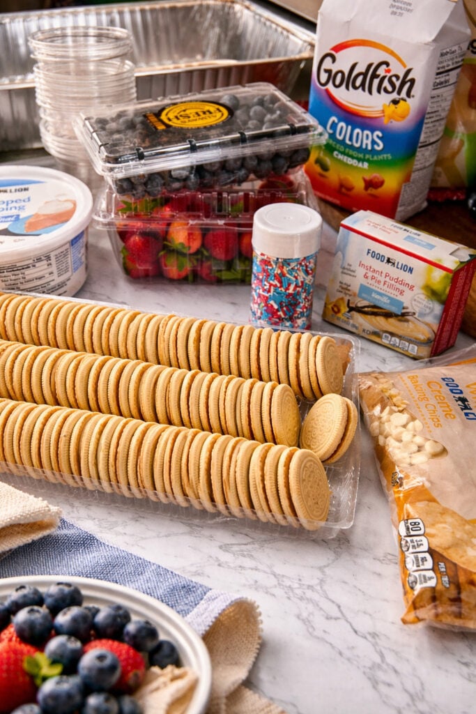 Ingredients for easy 4th of July vanilla dirt cups including Golden Oreos, instant pudding mix, Cool Whip, fresh berries, and festive sprinkles on kitchen counter