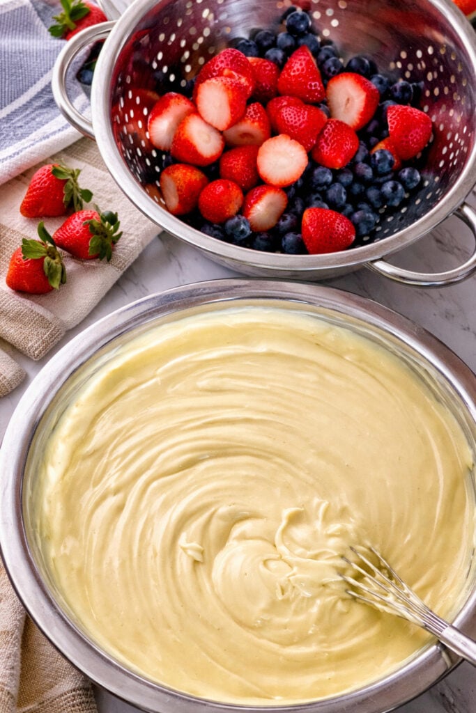 Creamy vanilla pudding mixture being whisked in a large bowl with fresh strawberries and blueberries nearby for easy 4th of July vanilla dirt cups