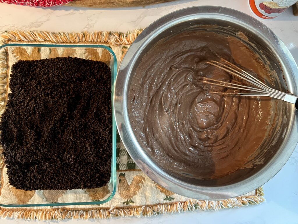Thick chocolate pudding mixture next to crushed Oreo cookie crumbs for worms in dirt dessert recipe, ready for layering into dirt pudding cups