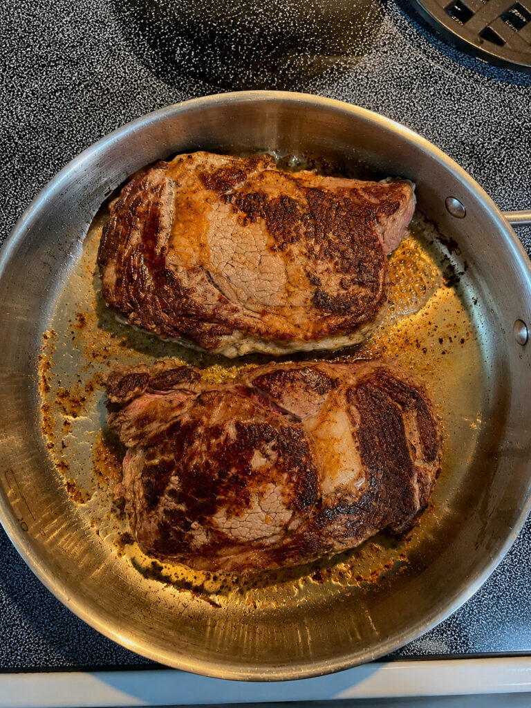 Perfectly seared ribeye steaks browning in a stainless steel skillet for steak and shrimp with parmesan sauce.