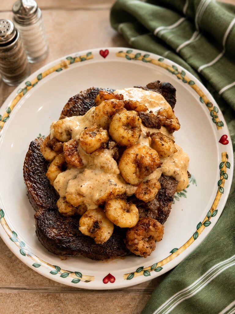 Ribeye steak topped with seasoned shrimp and creamy parmesan sauce served in a bowl with a green kitchen towel nearby.