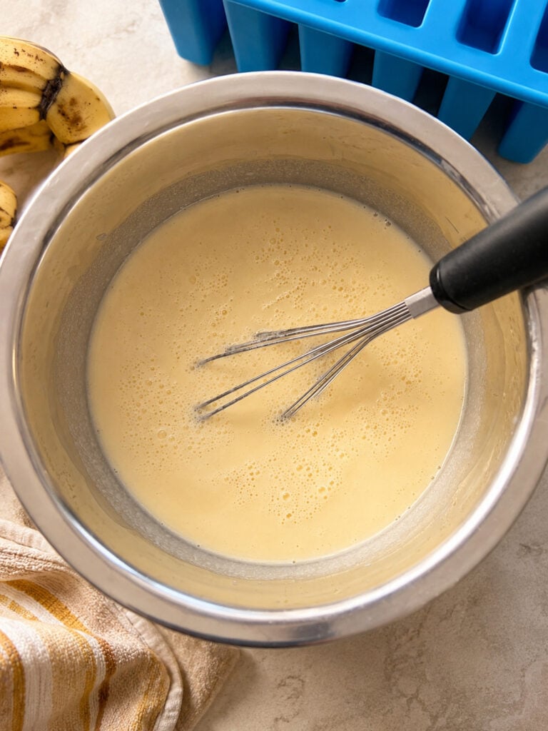 Smooth and creamy banana pudding mixture whisked in a metal mixing bowl with ripe bananas and popsicle molds in the background.