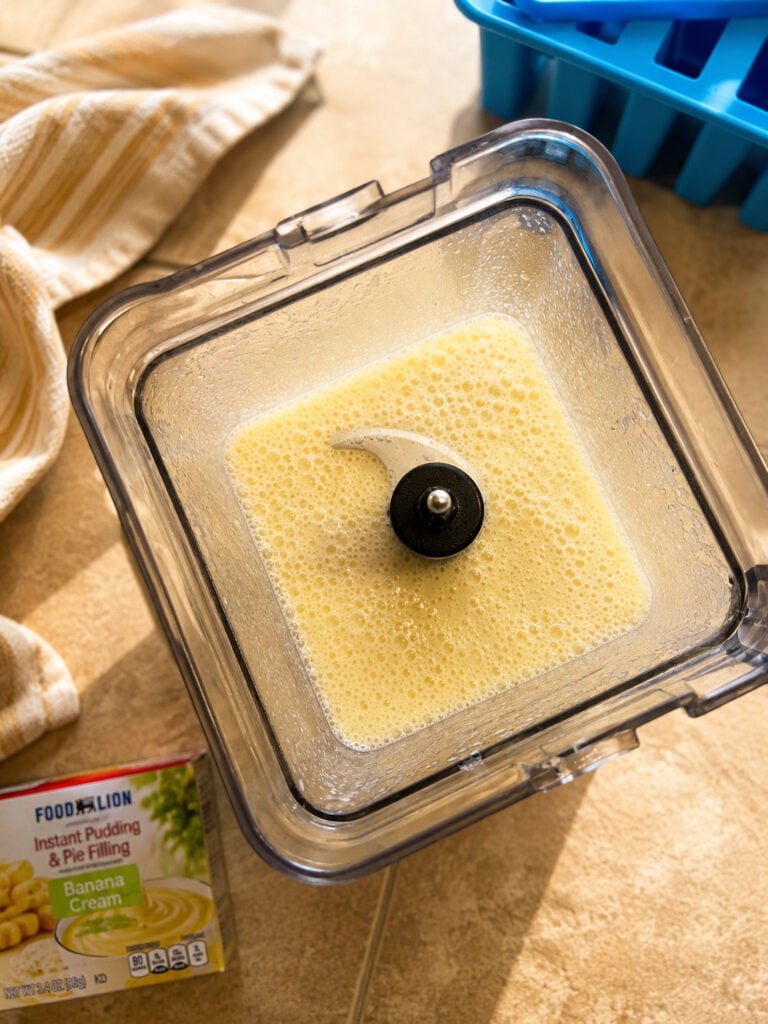 Creamy banana pudding mixture blended in a blender container beside a blue silicone popsicle mold and striped kitchen towel on a countertop.