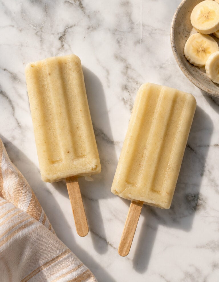 Three creamy homemade frozen banana treats with wooden sticks arranged on a marble surface beside fresh banana slices, ripe bananas, and striped kitchen towels.