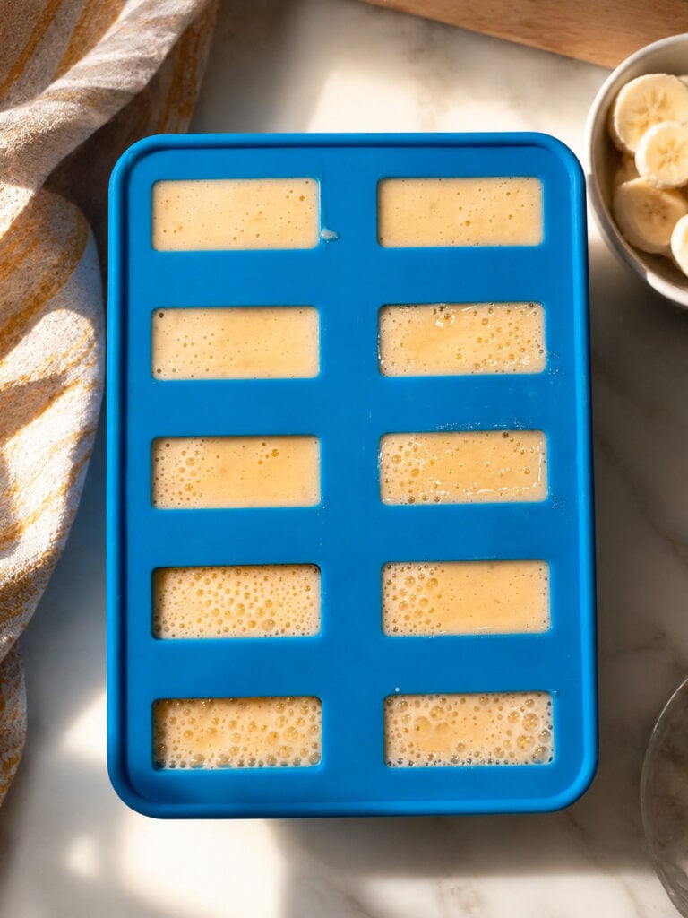 Blue silicone popsicle mold filled with creamy banana mixture before freezing on a marble countertop with sliced bananas nearby.
