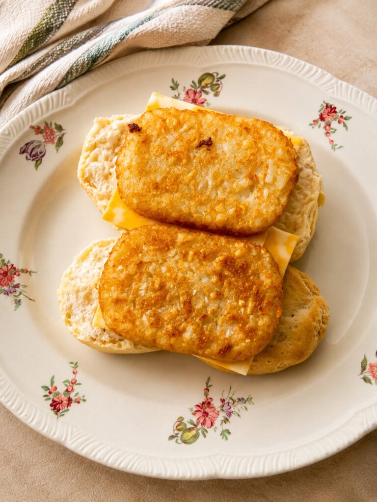 Split fluffy biscuit topped with sliced marbled cheese and crispy hash browns on a floral plate.