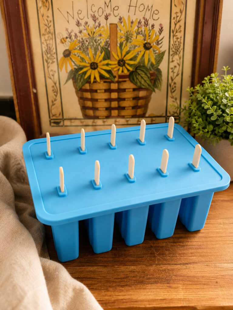 Blue silicone popsicle mold with lid and wooden sticks ready for freezing homemade chocolate pudding popsicles on a rustic wooden countertop.