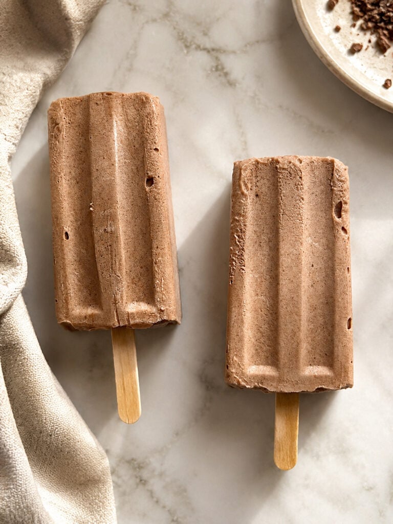 Two homemade chocolate popsicles on a marble surface with a soft kitchen towel and chocolate pieces, styled for a summer dessert recipe.